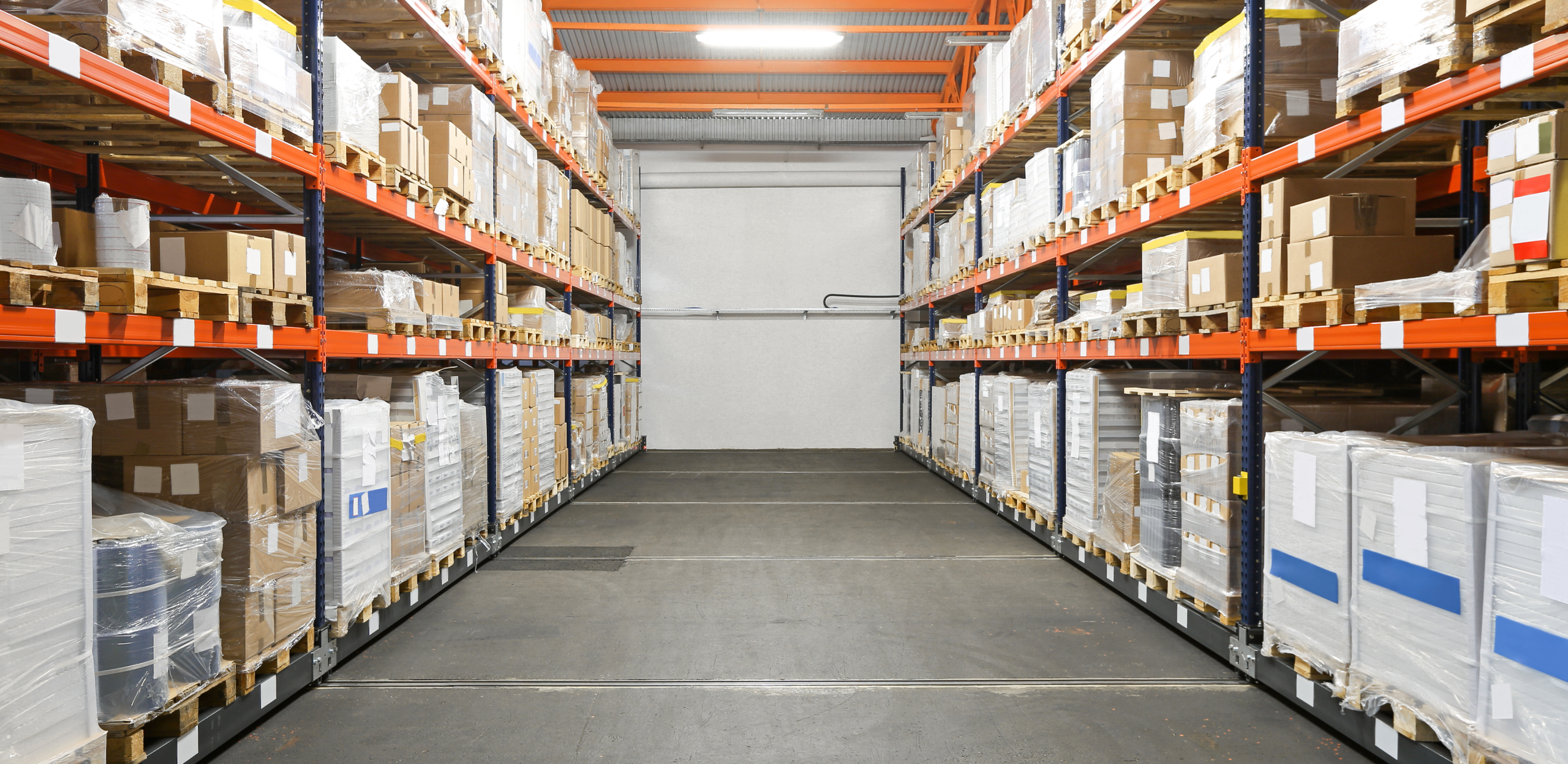 Modernes Hochregallager mit zwei langen Regalreihen, gefüllt mit palettierten Waren in Kartons und Folienverpackungen. Die Gänge sind sauber und ordentlich, die Regale stabil und in Orange-Blau gehalten. Optimale Bedingungen für Lagerung, Logistik und Kommissionierung.